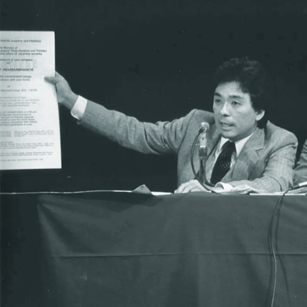 Frank Abe testifies at the Seattle CWRIC hearing, holding a poster from the first Day of Remembrance in 1978 — an early grassroots event that helped spark the redress movement. Photo: Densho / Frank Abe Collection
